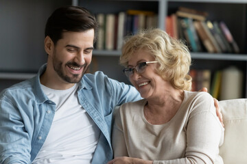 Wall Mural - Head shot happy young bearded man embracing beautiful smiling middle aged senior mother in eyewear, relaxing together on comfortable sofa in living room, enjoying funny conversation or gossiping.