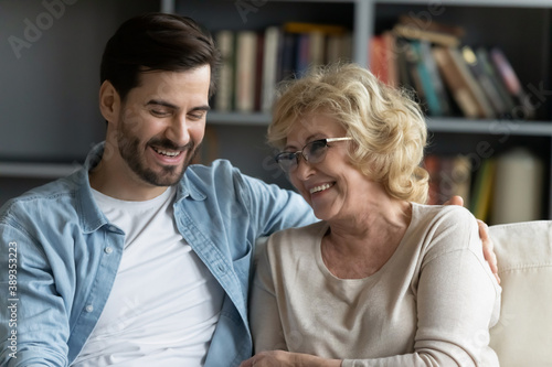 Cuadros en Lienzo Head shot happy young bearded man embracing beautiful smiling middle aged senior mother in eyewear, relaxing together on comfortable sofa in living room, enjoying funny conversation or gossiping
