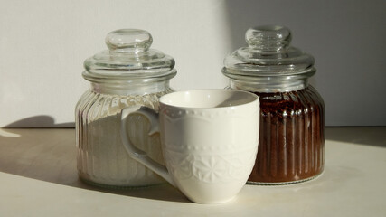 two glass bottles with ground coffee and sugar and a white ceramic cup on the table                               