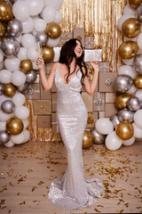 Wall Mural - birthday, Christmas or new year celebration concept - happy brunette woman in sexy silver dress with glass of champagne dancing over festive decorations of gift boxes and air balloons