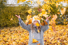 Young Girl Is Playing With Yellow Fallen Leaves And Smiling. Child Is Throwing Maple Leaves Up In Autumn Park. Autumn And Fall Concept.