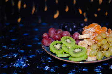 Fruits, grapes, tangerines, bananas, kiwi in a blue porcelain plate. black wooden floor, fruit plate