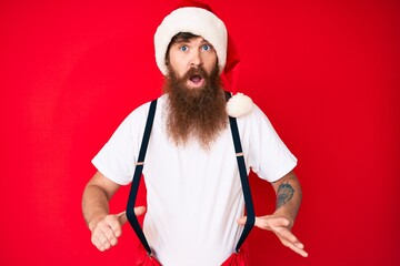 Wall Mural - Handsome young red head man with long beard wearing santa claus costume with suspenders clueless and confused expression. doubt concept.