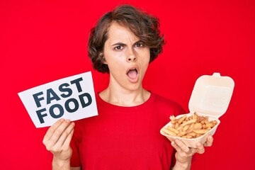 Poster - young hispanic woman holding potato chip and fast food banner in shock face, looking skeptical and s