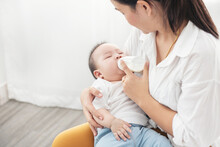 Portrait Of A Happy Asian Mother Giving Bottle Feeding To Her Baby In White Bedroom.  Baby Is Drinking Milk From A Bottle Hold By The Mother.