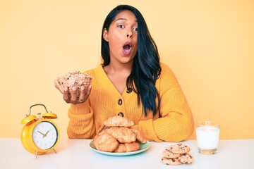 Canvas Print - Beautiful latin young woman with long hair sitting on the table having breakfast scared and amazed with open mouth for surprise, disbelief face