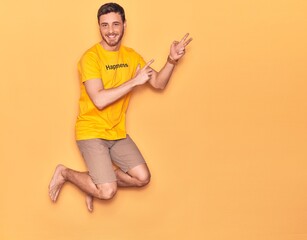 Wall Mural - Young handsome hispanic man wearing t shirt with happiness word message smiling happy. Jumping with smile on face over isolated yellow background