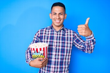 Young handsome latin man holding popcorn smiling happy and positive, thumb up doing excellent and approval sign