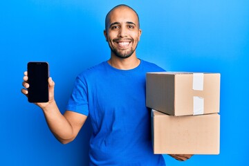 Sticker - Hispanic adult man holding delivery packages and showing smartphone screen smiling with a happy and cool smile on face. showing teeth.