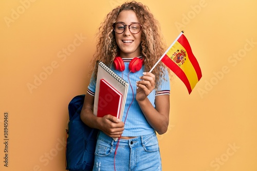 Beautiful caucasian teenager girl exchange student holding spanish flag winking looking at the camera with sexy expression, cheerful and happy face.