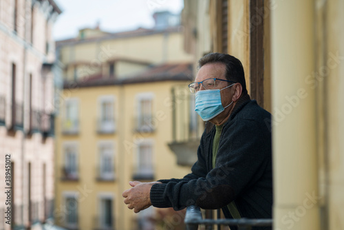 mature man in balcony home lockdown during covid19 outbreak - senior male on his 70s in face mask worried and tired looking to the street thoughtful and depressed in quarantine