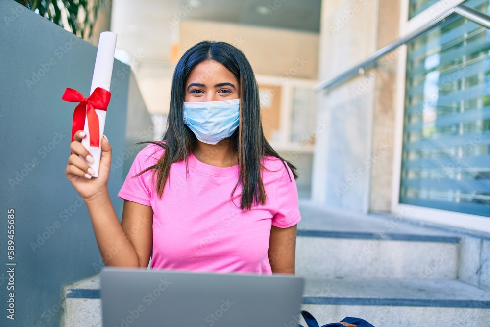 Young latin student girl wearing medical mask using laptop and holding diploma at university.
