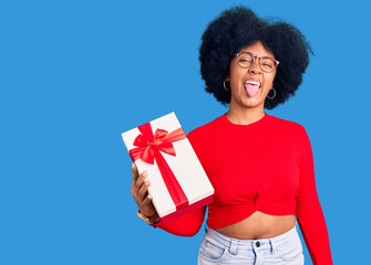 Wall Mural - Young african american girl holding gift sticking tongue out happy with funny expression. emotion concept.