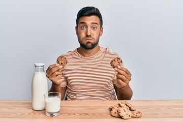 Poster - Handsome hispanic man drinking a glass of milk holding chocolate cookies puffing cheeks with funny face. mouth inflated with air, catching air.