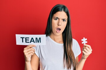 Poster - Young brunette woman holding team paper and piece of puzzle as teamwork in shock face, looking skeptical and sarcastic, surprised with open mouth