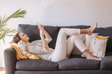 Young woman lying on couch and texting talking at social media on smartphone. Middle age woman on sofa typing messaging on mobile phone. Chat talk with friends online on the Internet.