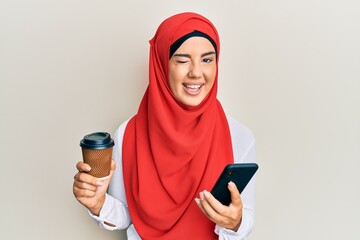 Wall Mural - Young beautiful hispanic girl wearing islamic hijab drinking a coffee usign smartphone winking looking at the camera with sexy expression, cheerful and happy face.