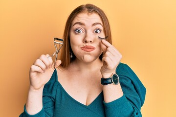 Sticker - Young beautiful redhead woman holding eyelash curler and fake lashes puffing cheeks with funny face. mouth inflated with air, catching air.