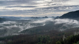 Fototapeta Las - Panoramablick in den Morgenstunden von den Affensteinen zum Kleinen Winterberg. Die Sonne schickt ihre ersten wärmenden Strahlen durch den dichten Nebel in die Bergwelt des Nationalparkes.