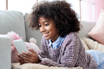 Happy african american child kid girl holding phone relaxing on couch. Cute mixed race child using social media mobile apps on smartphone tech, watching videos on cellphone lying on sofa at home.