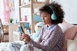© insta_photos - African american child kid girl wearing headphones holding phone sitting on bed. Cute black kid using smartphone tech listening course watching video class elearning on cellphone in bedroom at home.