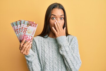 Sticker - Beautiful hispanic woman holding 100 new zealand dollars banknote covering mouth with hand, shocked and afraid for mistake. surprised expression