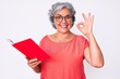 © Krakenimages.com - Senior hispanic woman wearing glasses holding book doing ok sign with fingers, smiling friendly gesturing excellent symbol