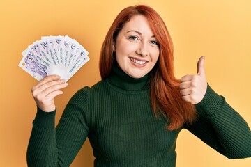 Beautiful redhead woman holding 2 singapore dollars banknotes smiling happy and positive, thumb up doing excellent and approval sign