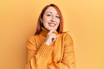 Poster - Beautiful redhead woman wearing casual winter sweater over yellow background looking confident at the camera smiling with crossed arms and hand raised on chin. thinking positive.