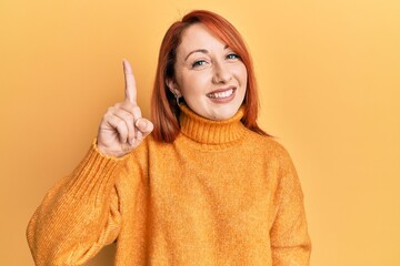 Poster - Beautiful redhead woman wearing casual winter sweater over yellow background pointing finger up with successful idea. exited and happy. number one.