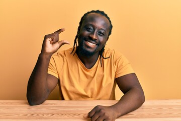 Sticker - Handsome young black man wearing casual clothes sitting on the table smiling and confident gesturing with hand doing small size sign with fingers looking and the camera. measure concept.