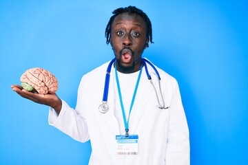 Poster - Young african american man with braids wearing doctor coat holding brain scared and amazed with open mouth for surprise, disbelief face