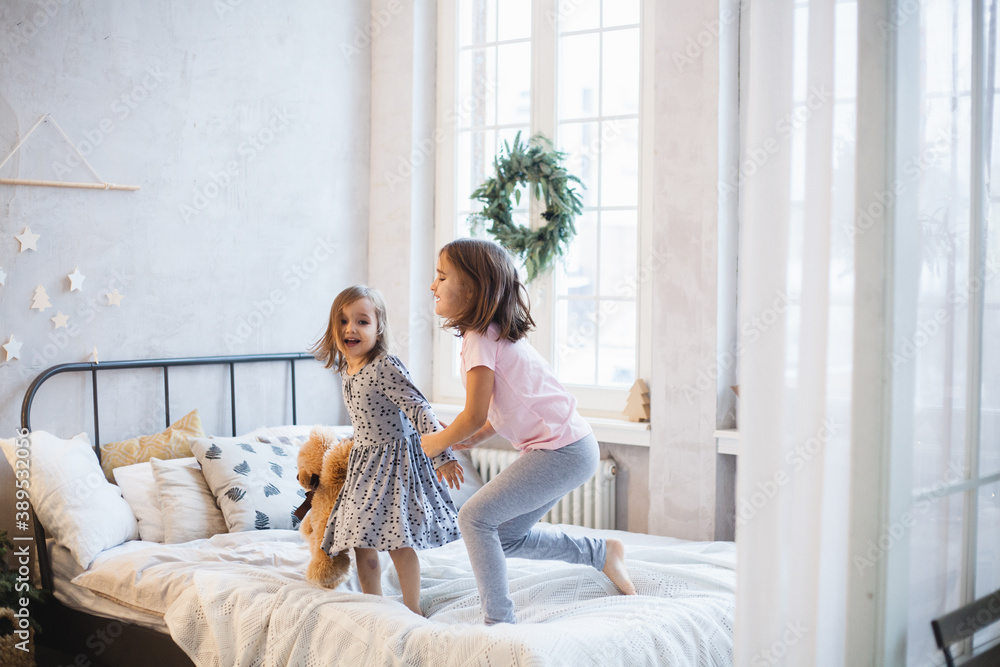 The room is decorated for the new year, the little sisters are sitting on the bed, playing games, pillows, laughing, waiting for Christmas