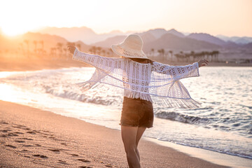 Wall Mural - A woman in a hat walks at sunset on the beach rear view