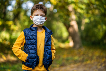 Portrait of young boy in park in autumn. He is wearing protective face mask. Covid-19 pandemic concept.