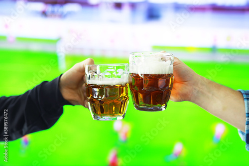 Two hands horizontal holding lager beer glass and clinking on background of football game. Sport fans cheer up. Friends leisure lifestyle concept.