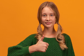 Image of charming blonde teen smiling and showing thumb up. Studio shot, yellow background. Facial expression concept