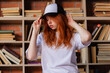 © oleg_ermak - Portrait of a redhead woman in template blank t-shirt and cap with healthy freckled skin. Young caucasian model posing on bookshelf background. Copy space and mock up. Place for adverising