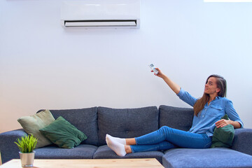 Wall Mural - Young happy woman sitting on couch under air conditioner and adjusting comfort temperature with remote control at modern home