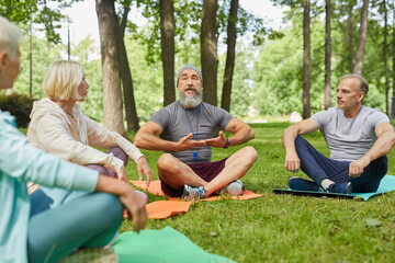 Stylish bearded yoga trainer sitting on mat demonstrating his senior clients how to do breath exercise