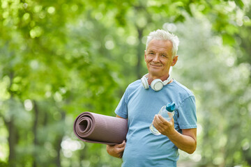 Waist up portrait shot of active senior man holding yoga mat and bottle of water looking at camera