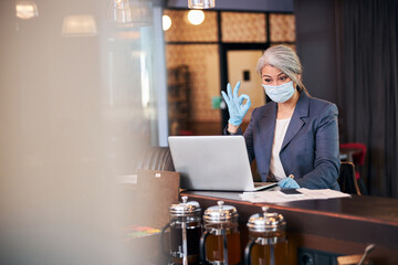 Wall Mural - Businesswoman in medical mask using laptop and showing ok gesture