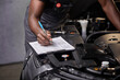 © alfa27 - african professional auto service technician in uniform standing near car hood repairing and using check list for car inspect, indoors