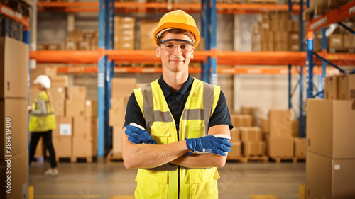 Handsome Smiling Worker Wearing Hard Hat, Standing with Crossed Arms in the Retail Warehouse full of Shelves with Cardboard Boxes. Professional Working in Logistics, Delivery, and Distribution Center