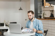 © ViDi Studio - Serious handsome young man sitting alone at table in coffee shop cafe restaurant indoors working or studying on laptop pc computer with papers document. Freelance mobile office business concept.