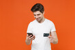© ViDi Studio - Smiling cheerful funny handsome young man 20s wearing basic casual empty blank white t-shirt using mobile cell phone hold credit bank card isolated on bright orange colour background studio portrait.