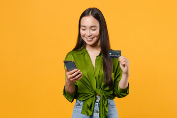 Wall Mural - Smiling cheerful young brunette asian woman wearing basic green shirt standing hold in hands using mobile cell phone hold credit bank card isolated on bright yellow colour background, studio portrait.