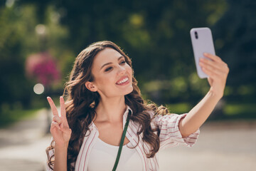 Wall Mural - Photo of sweet cheerful long curly hair young lady doing selfie show v-sign enjoy weekend in park wear striped shirt outside