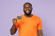© ViDi Studio - Smiling funny young african american man 20s wearing basic casual orange blank empty t-shirt standing hold credit bank card looking camera isolated on pastel violet colour background studio portrait.