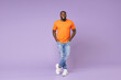 © ViDi Studio - Full length side view of smiling young african american man 20s wearing basic casual orange t-shirt holding hands in pockets looking camera isolated on pastel violet colour background studio portrait.